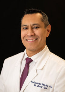 A man wearing a white coat embroidered with Professional Hearing Aid Dr. David Jara, a white shirt, and a maroon tie, smiling against a dark background.