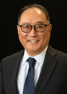 A man wearing glasses, a dark suit, a white dress shirt, and a navy tie smiles in front of a black background.
